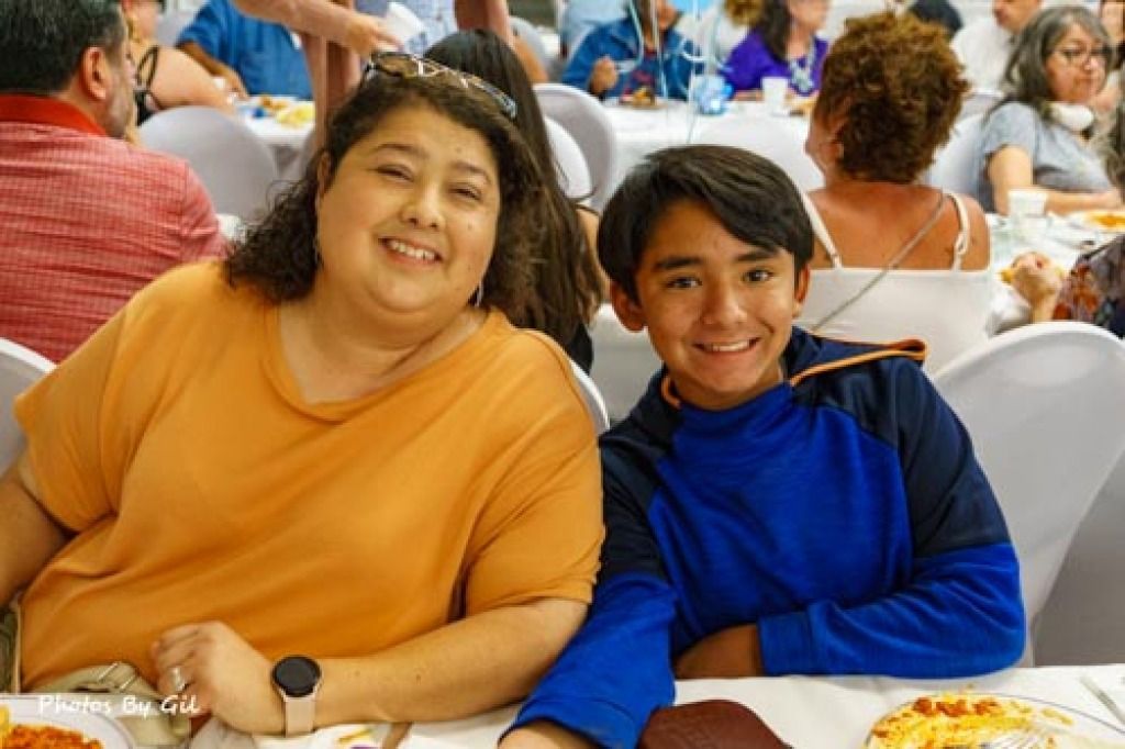 A woman in an orange shirt and a boy in a blue hoodie smile at a table during a lively gathering.