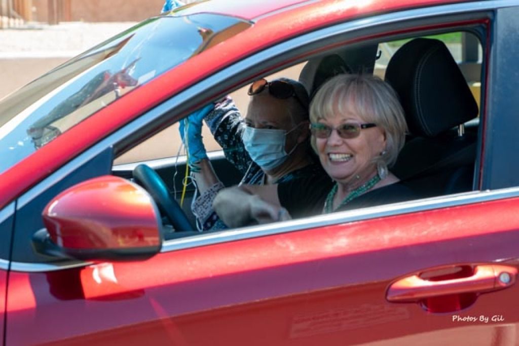 A smiling woman drives a red car while a passenger in a mask raises a hand. 