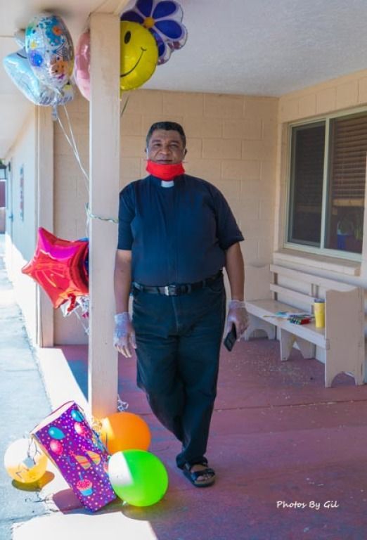 A man in a clerical shirt and mask stands outside, holding a phone. 