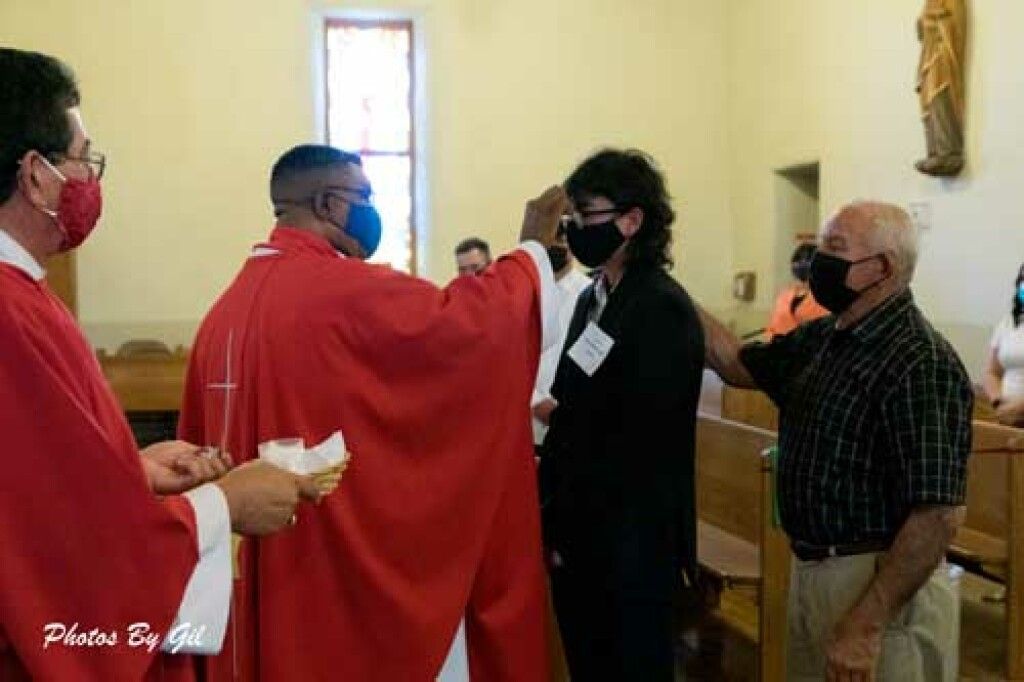 A priest in red vestments and a blue mask performs a blessing on a masked individual in a black suit inside a church. 
