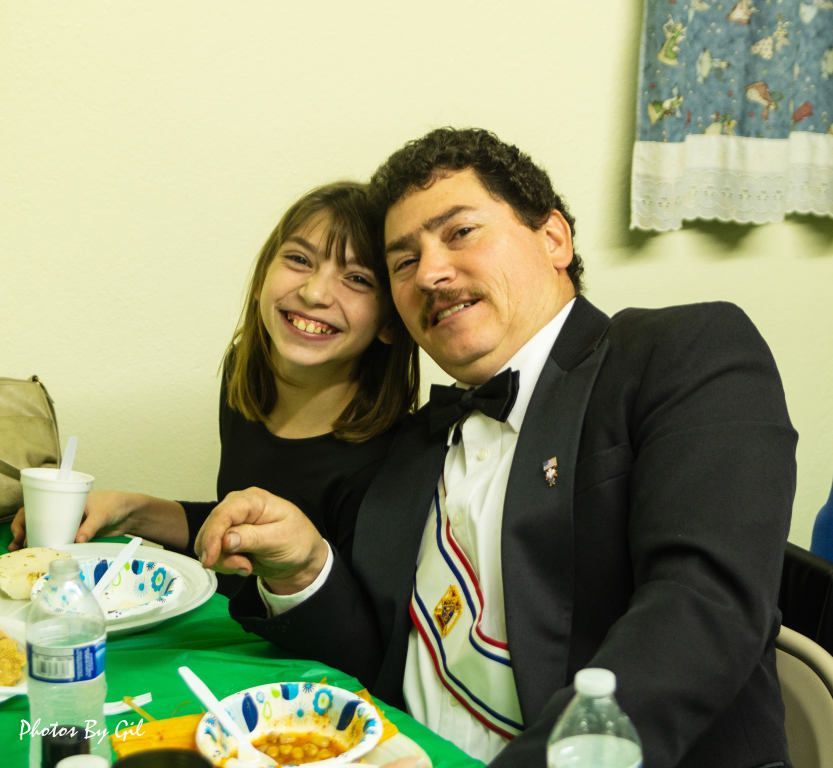 Man in a tuxedo and young girl smiling at a dining table with paper plates and bottled water.