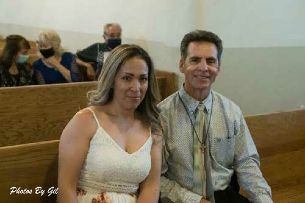 A woman in a white dress sits beside a man in a tie, smiling in a church pew.