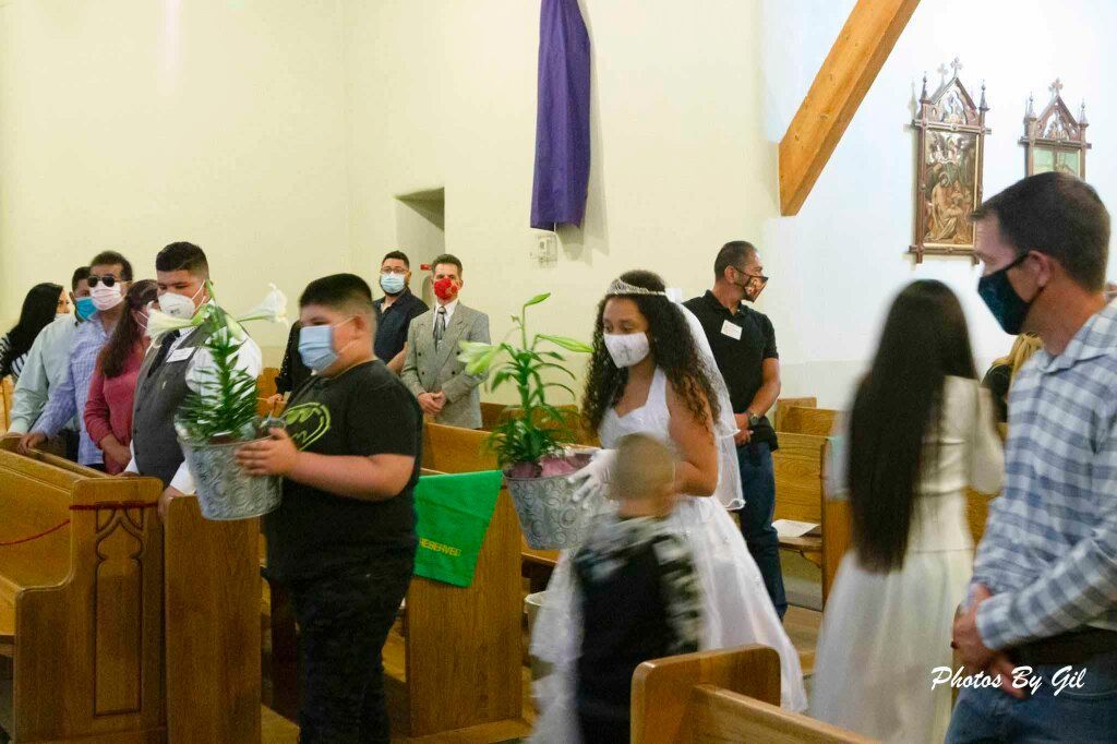 A group of masked people, including a child in a white dress carrying plants, process down a church aisle.