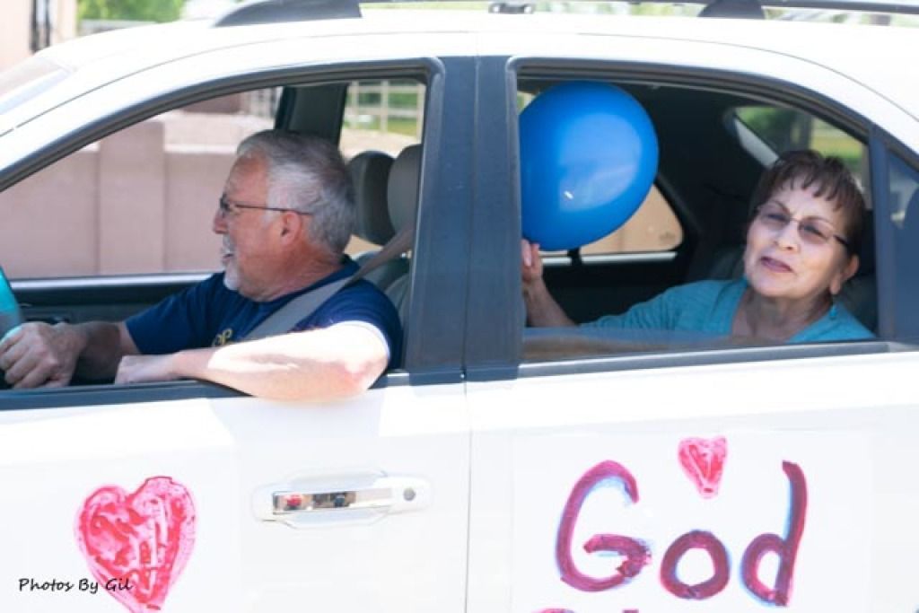 An elderly couple drives in a decorated white car, the man at the wheel, while the woman holds a blue balloon. 
