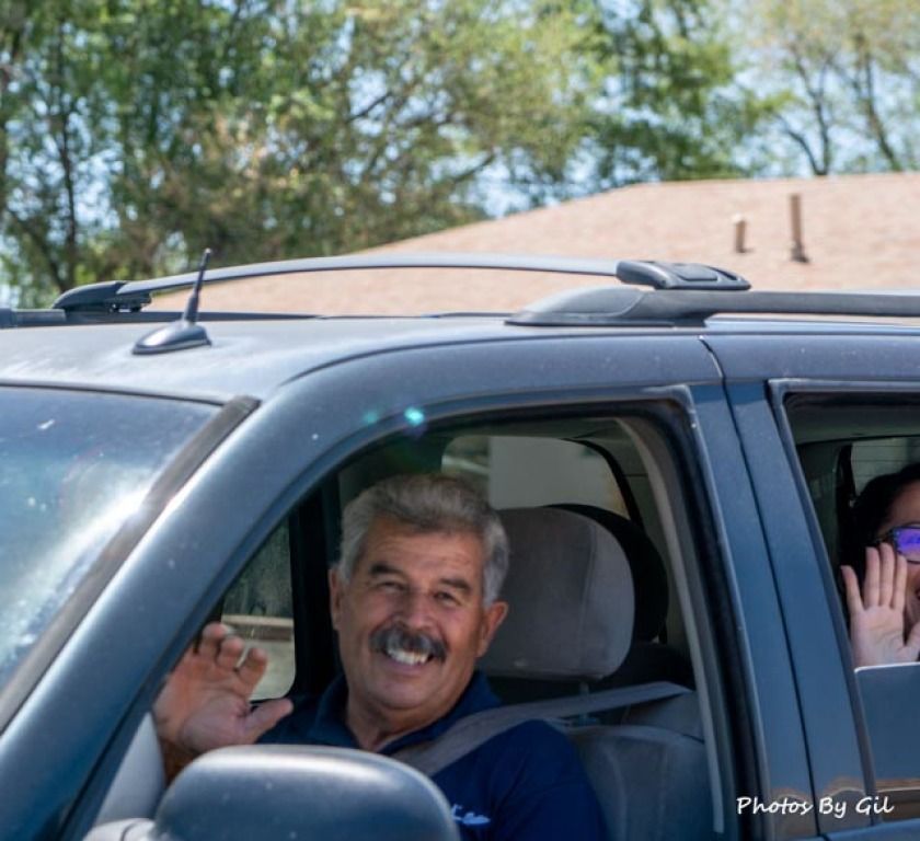 A smiling man with a mustache waves from the driver's seat of a blue SUV. 