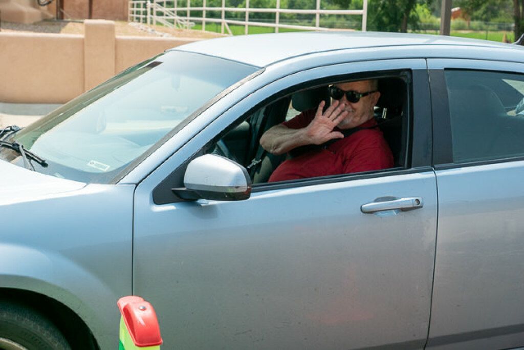 A man in sunglasses waves from the driver's seat of a parked silver car.