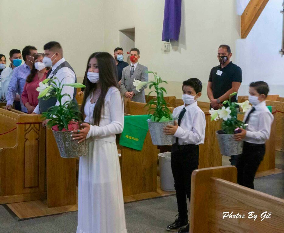 Young girl in a white dress and two boys in white shirts, all wearing masks, carry potted lilies in a church. 