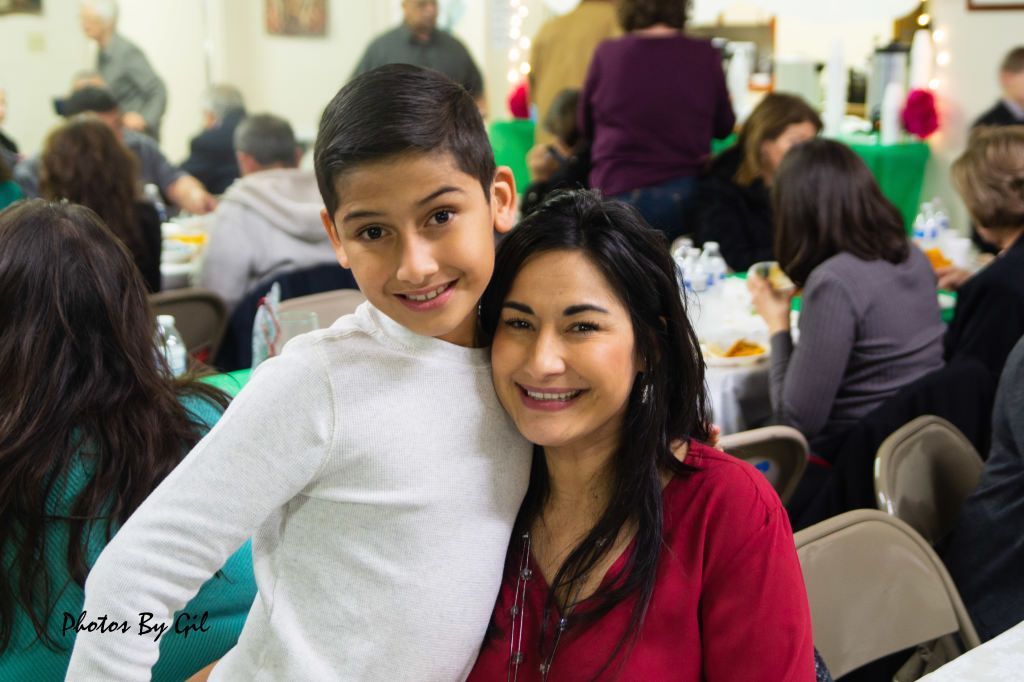 A smiling woman in a red blouse poses with a young boy in a white shirt. 