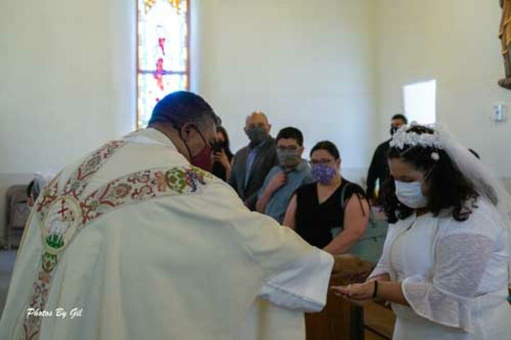 A priest, wearing ornate vestments, performs a blessing over a young woman dressed in white with a veil in a church.