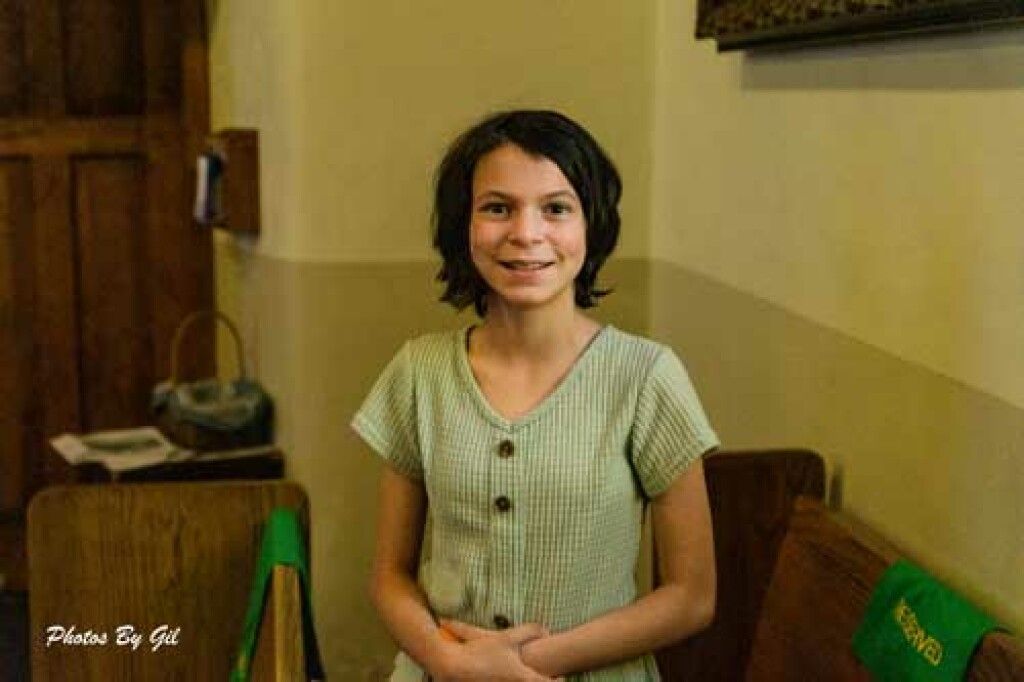 Smiling girl with short brown hair in a light green dress stands in a warmly lit room. 