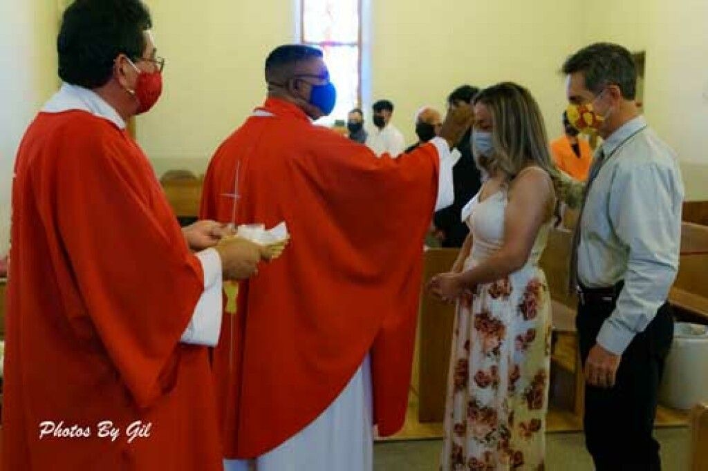 A priest in red robes blesses a woman wearing a floral dress, with a man beside her, in a church.