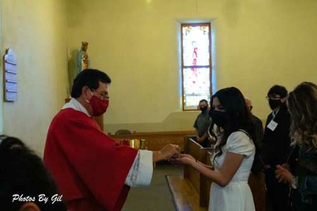 A priest in red vestments gives communion to a woman in white during a church service.