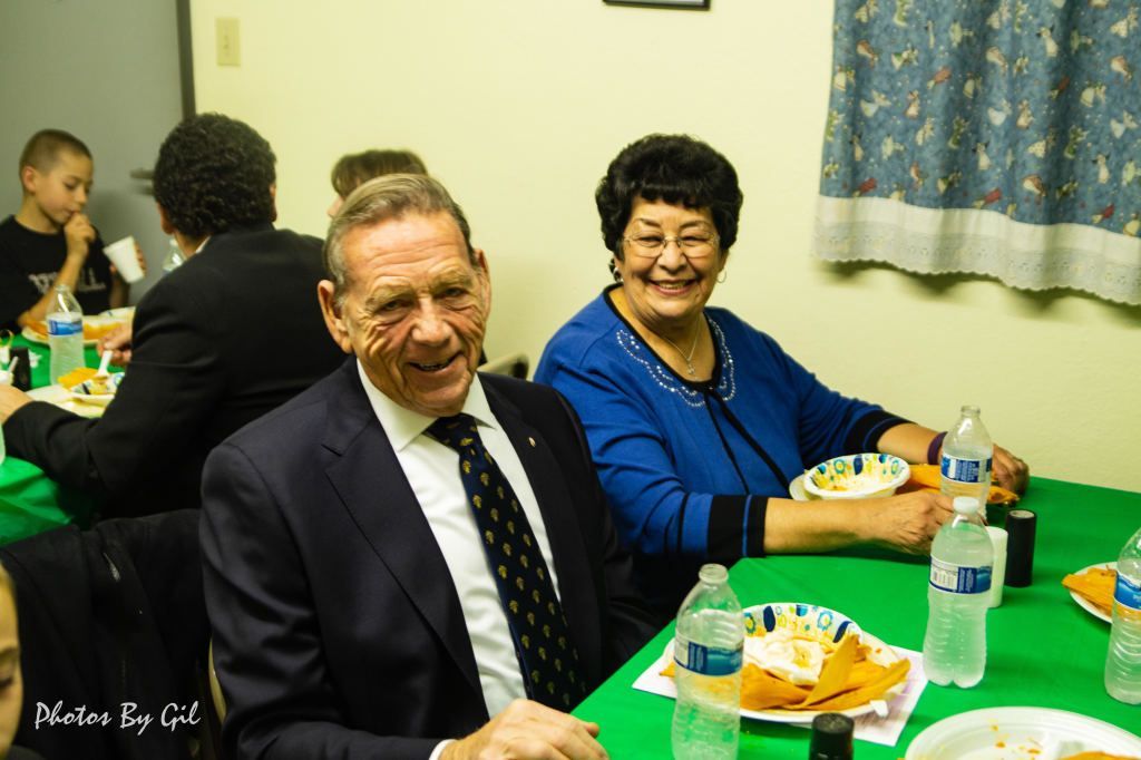 A smiling elderly couple sits at a green table set with paper plates and water bottles.