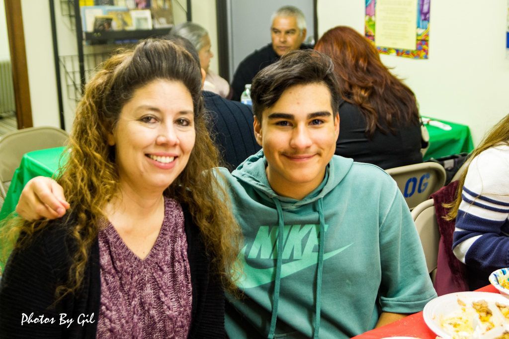 A smiling woman with wavy hair and a maroon sweater sits beside a young man in a green hoodie at a dining table.