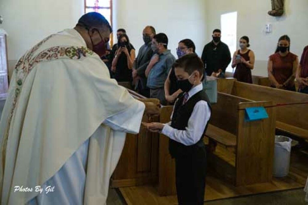A priest in a white robe gives communion to a young boy in a suit at a church.