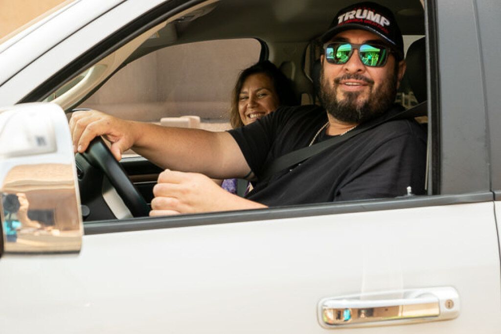 A smiling man wearing sunglasses and a baseball cap is driving a white vehicle.