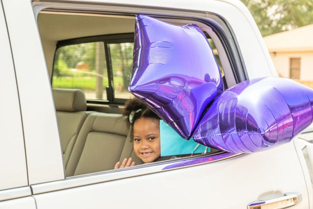A smiling child waves from a car window, with shiny purple star balloons tied to the door.