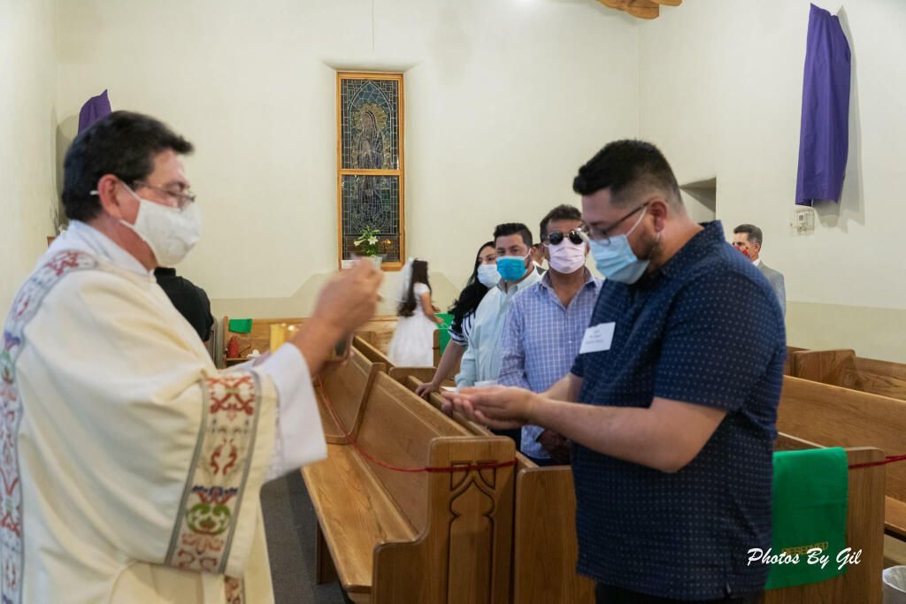 A priest in a white robe gives communion to a masked man during a church service. 