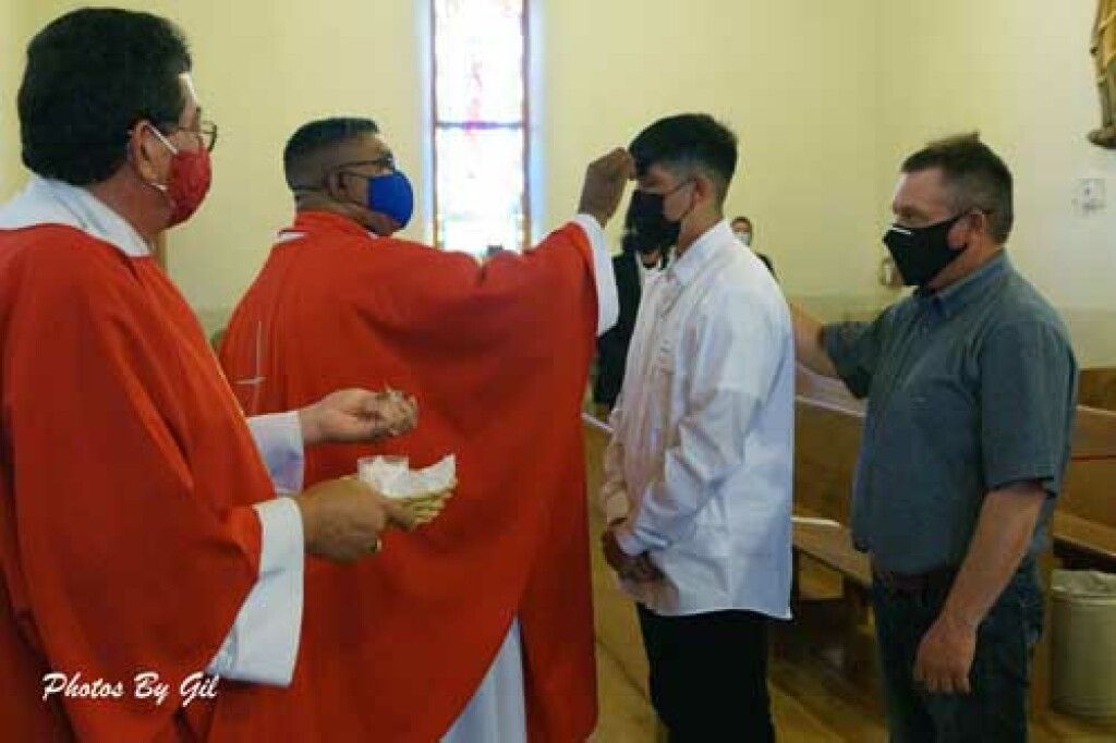 Priest in red robe blesses a young man wearing a white shirt and black mask during a church ceremony. 