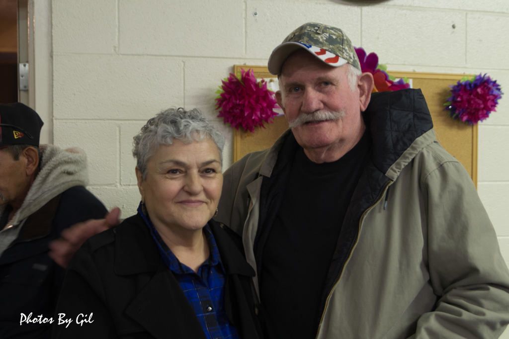 A smiling elderly couple stands in a room with colorful flowers on the wall.