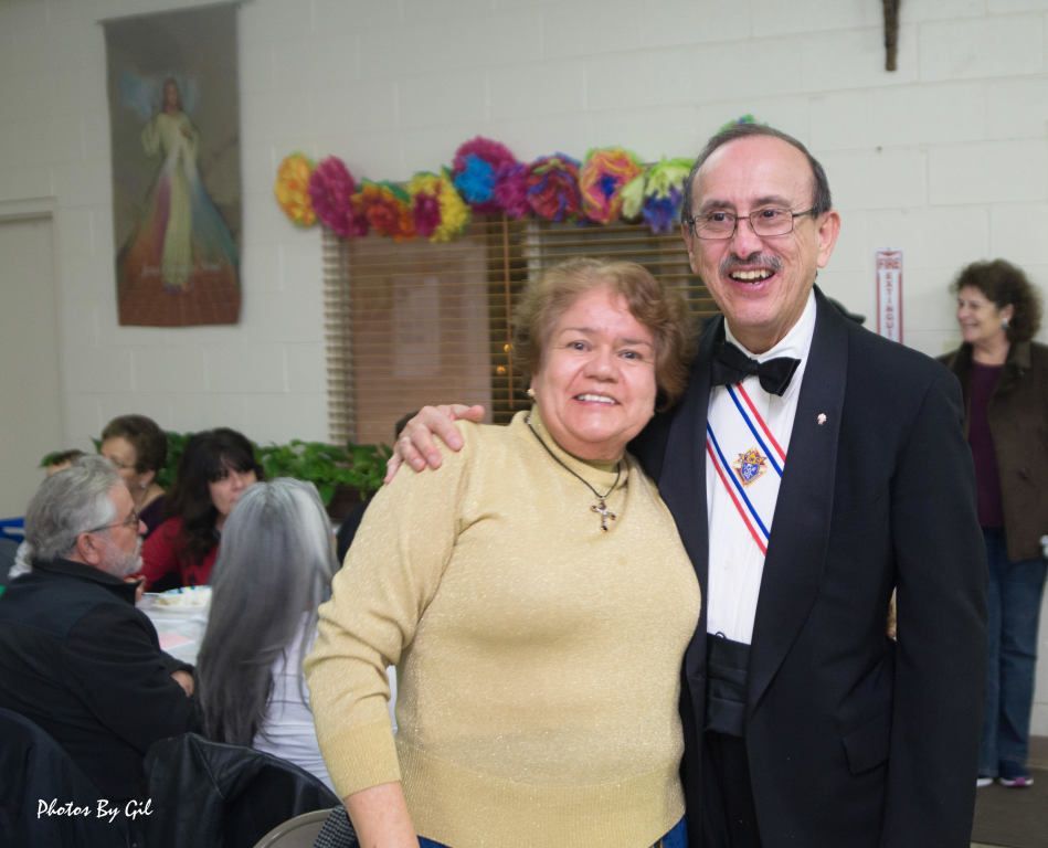 A smiling woman in a beige sweater and a man in a tuxedo with a medal.