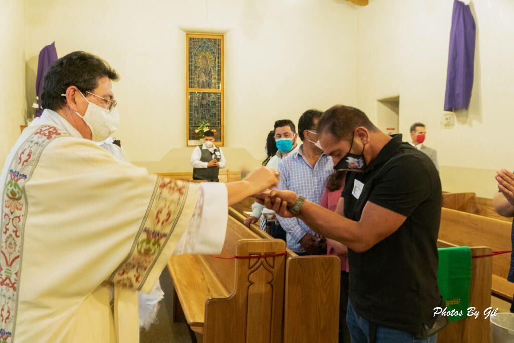 A priest in a white robe and mask offers a blessing to a kneeling man in a church. 