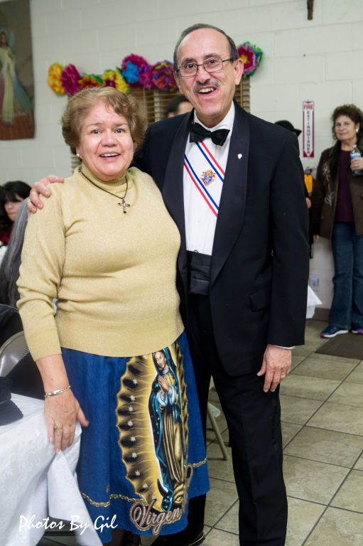 A smiling man in a tuxedo with a sash stands beside a woman in a beige sweater and skirt featuring the Virgin Mary. 