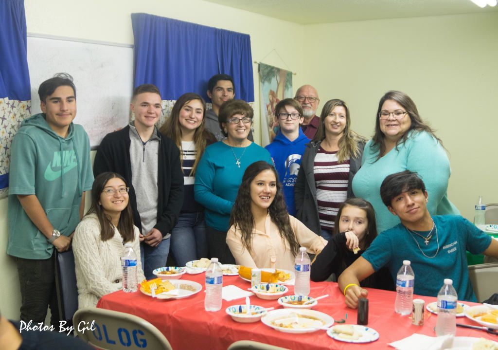 A group of smiling people is gathered in a room, some standing and others seated at a table with food and drinks.