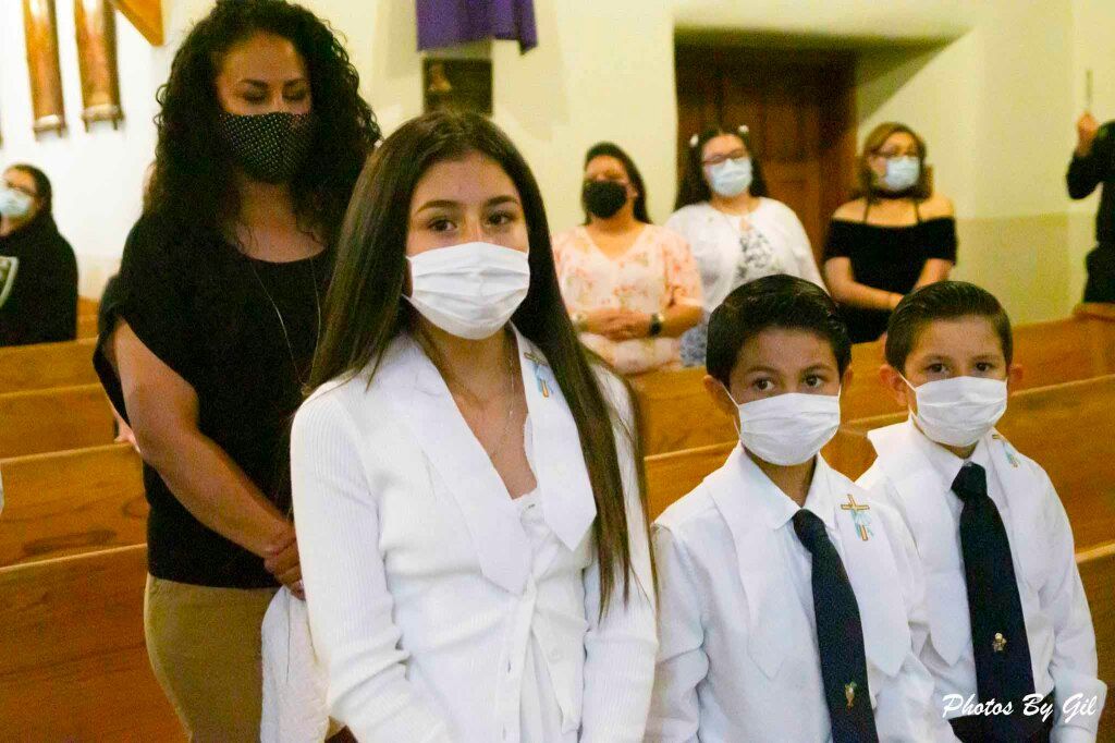 A young girl and two boys, all in white attire with ties and wearing masks, stand indoors.