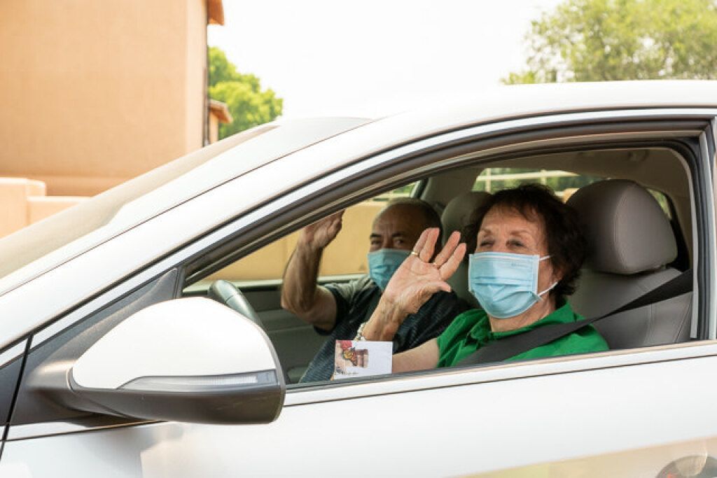 An older couple wearing masks waves from inside a parked car. 