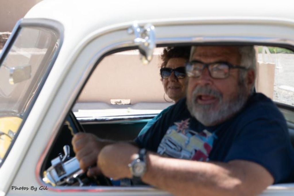 An older man with a beard and glasses is driving a classic car, looking surprised. 