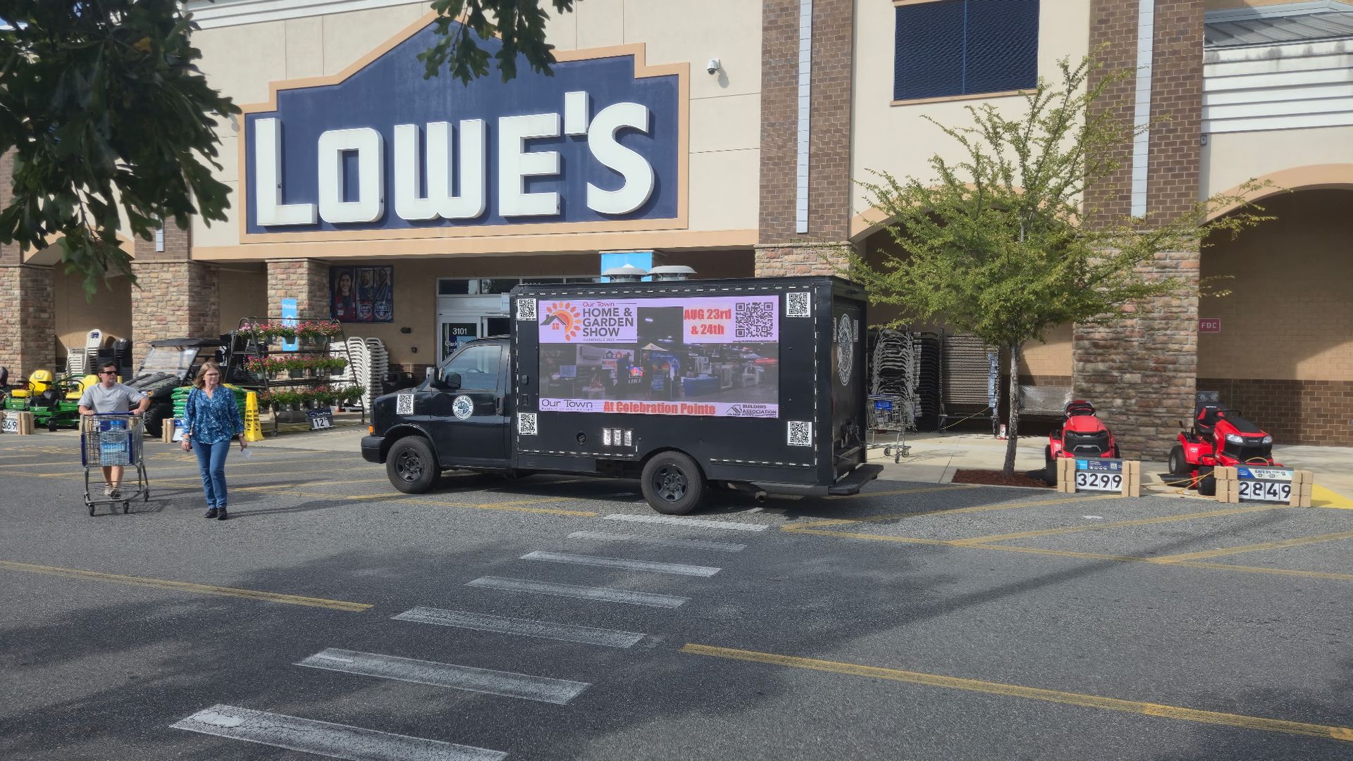 A black truck with a screen parked in front of Lowe's. People walk by.