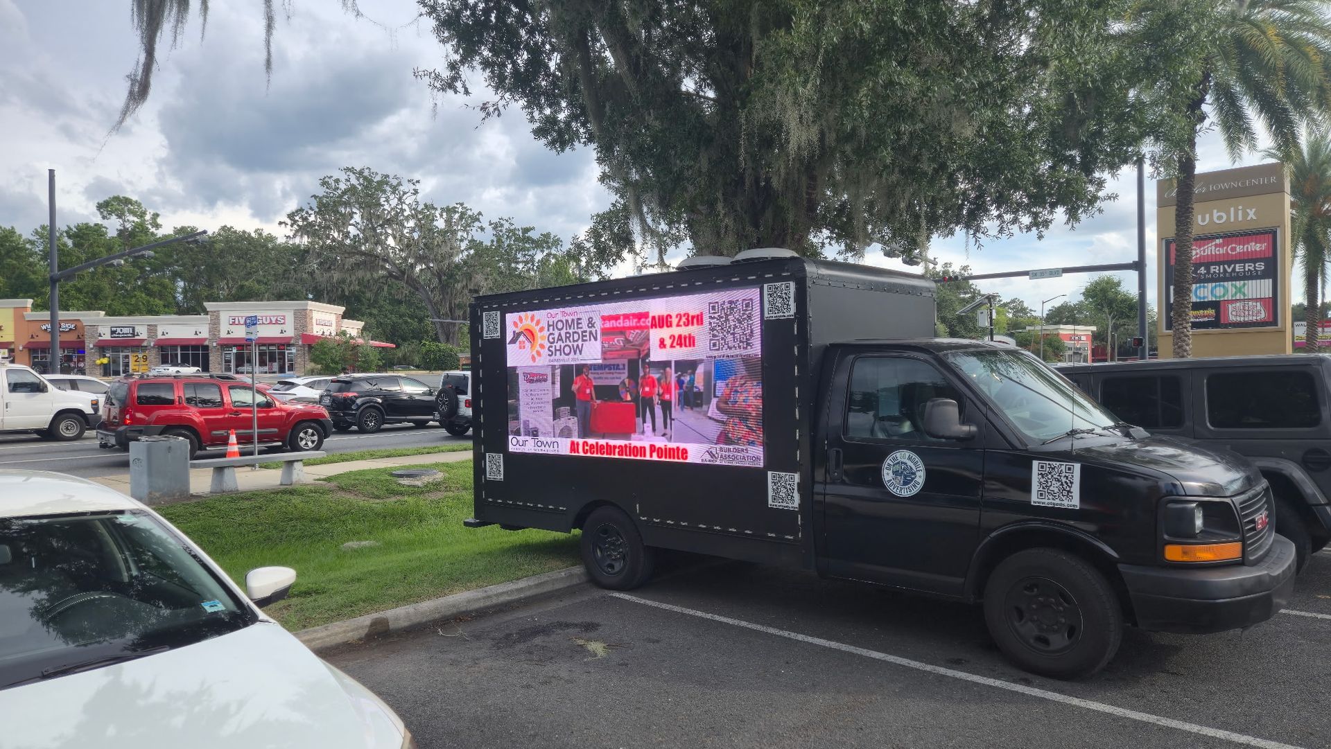 Black truck with an LED display parked in a lot. Shows advertisements. Buildings and cars in background.