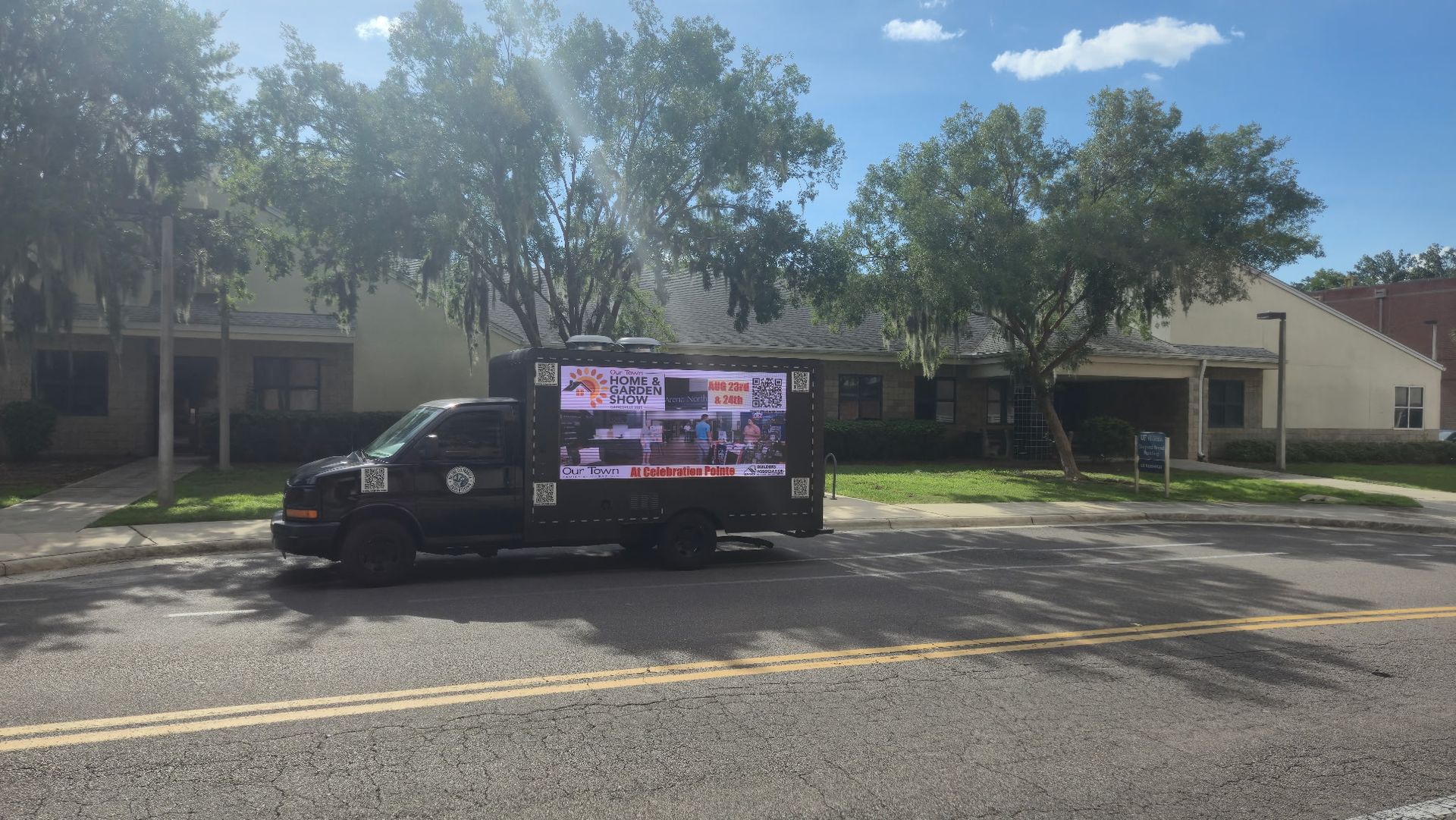 Black truck with digital screen parked on a street, showing videos, in front of houses on a sunny day.