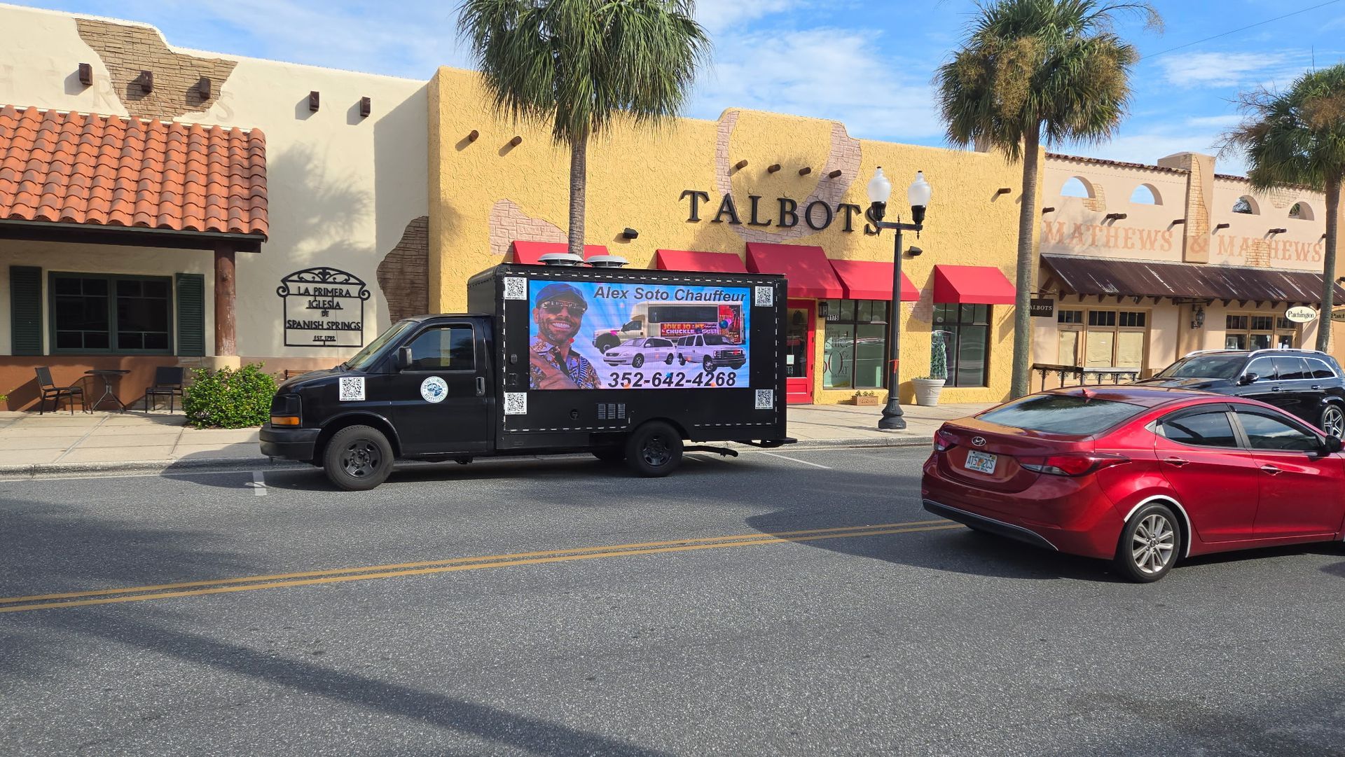 Black truck with digital display advertising on a street in front of a Talbots store, red car next to it.