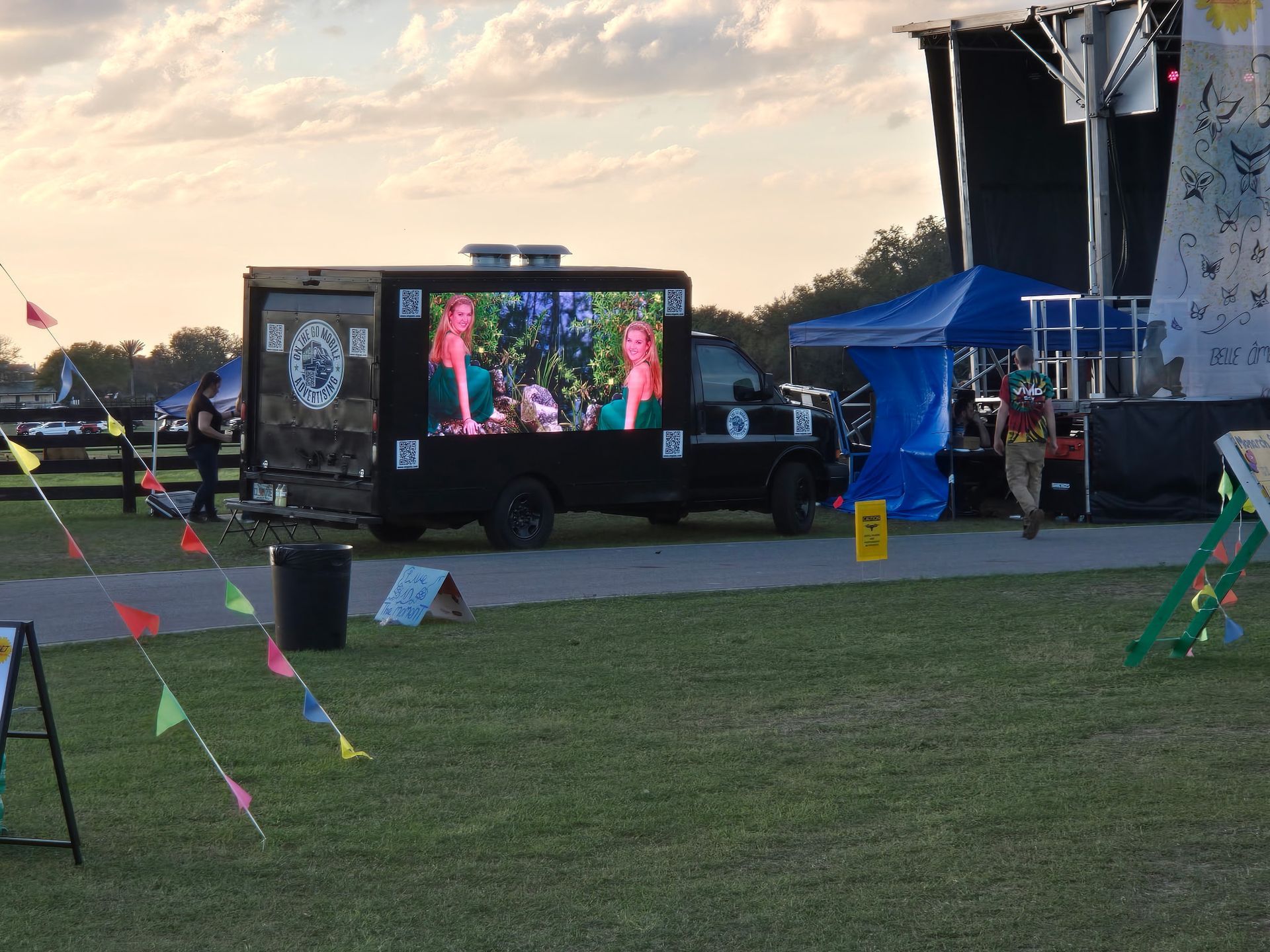 Black truck with large screen displaying video at outdoor event. Person walks by blue tent, flags strung nearby.