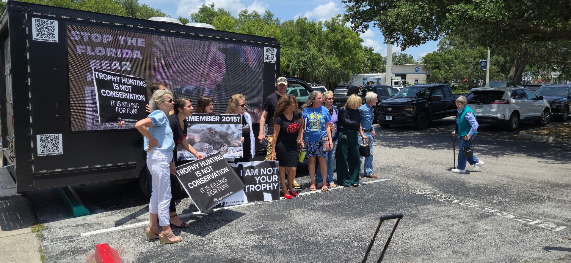 Protesters with signs stand near a truck with a screen displaying text. Cars and trees are in the background.