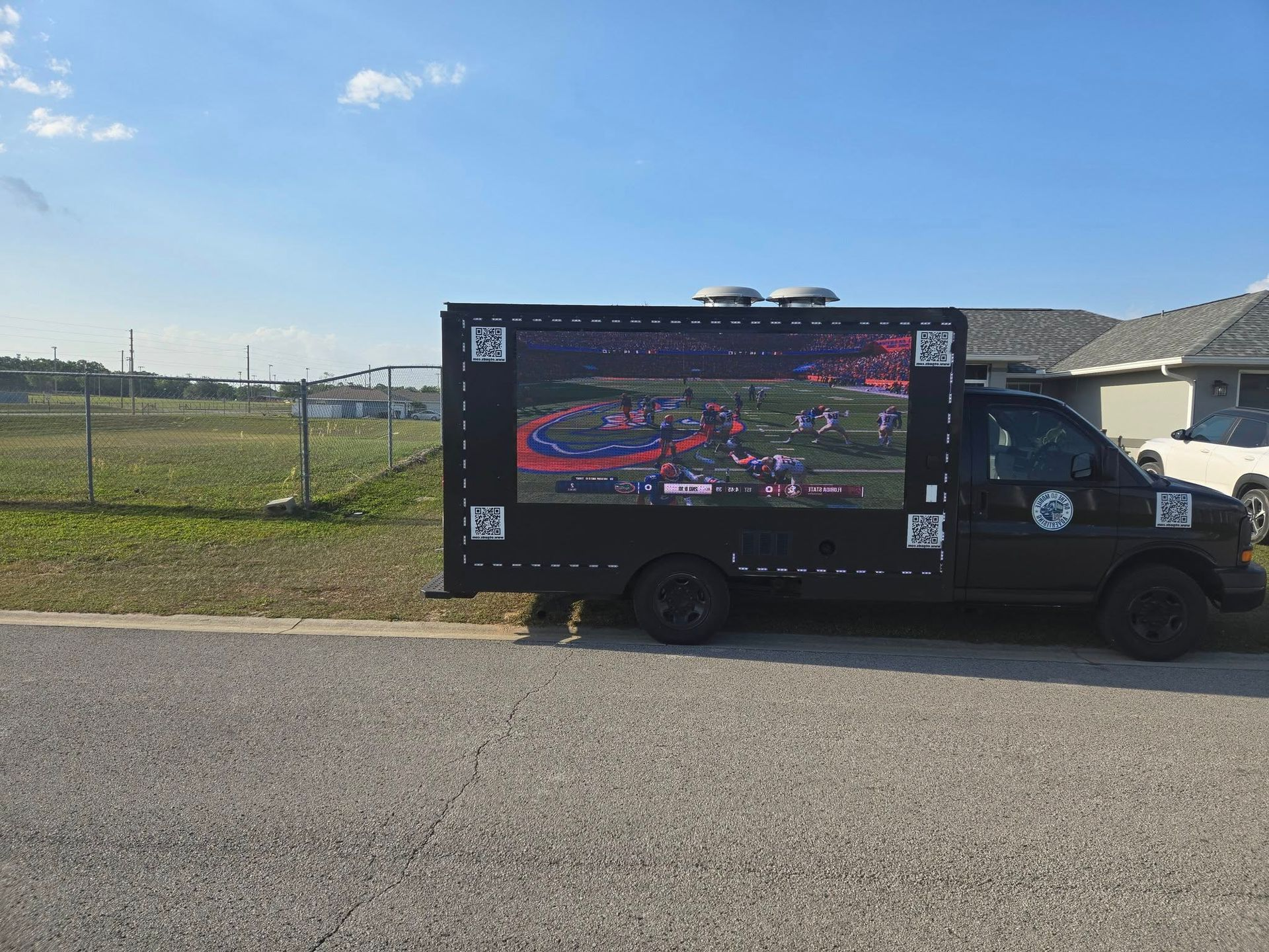 Black truck with large screen showing a sports game, parked on a road near a grassy field, houses in background.
