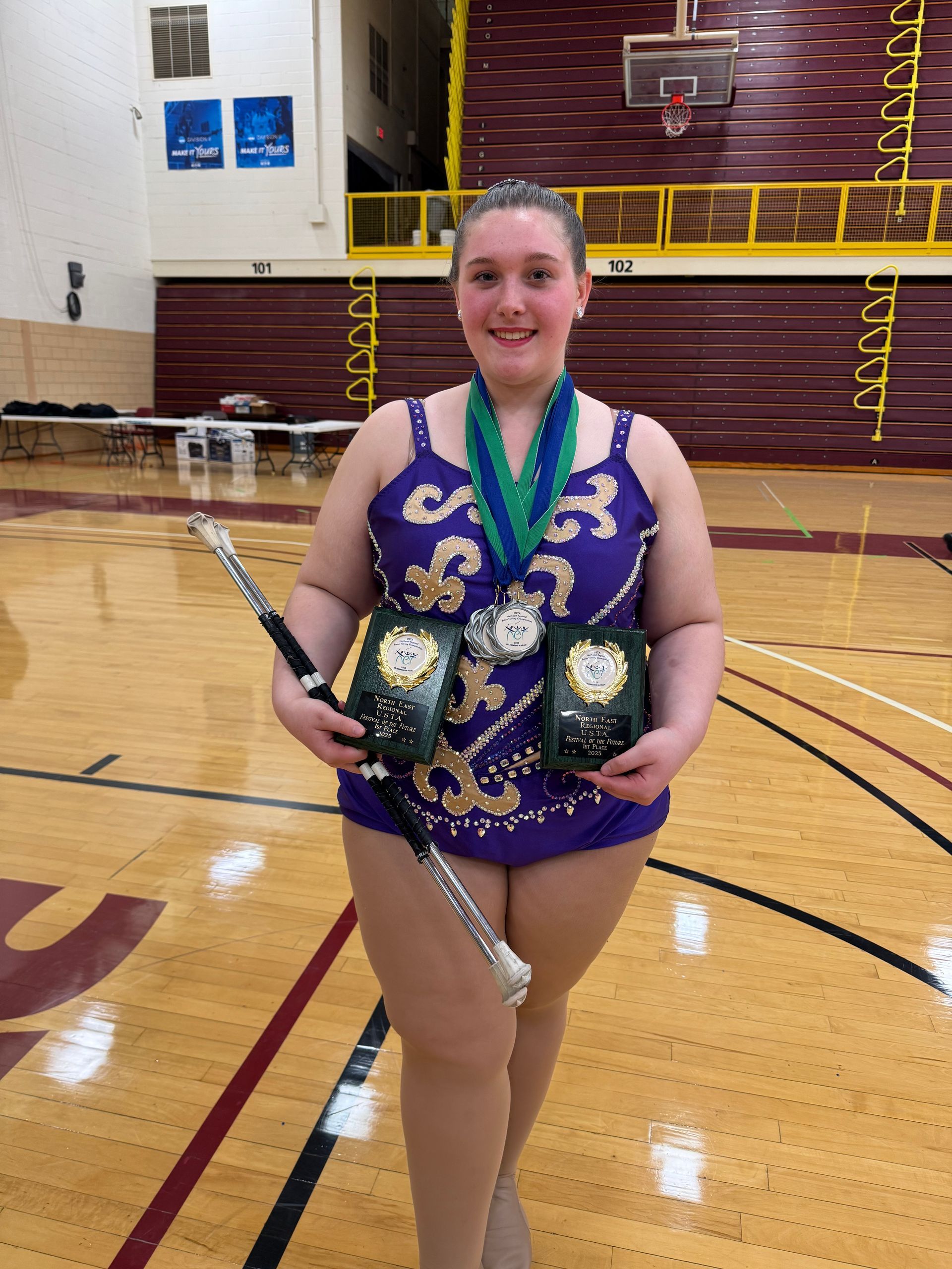 Young person in baton twirling uniform poses outside, smiling, holding baton. Bright colors on costume.