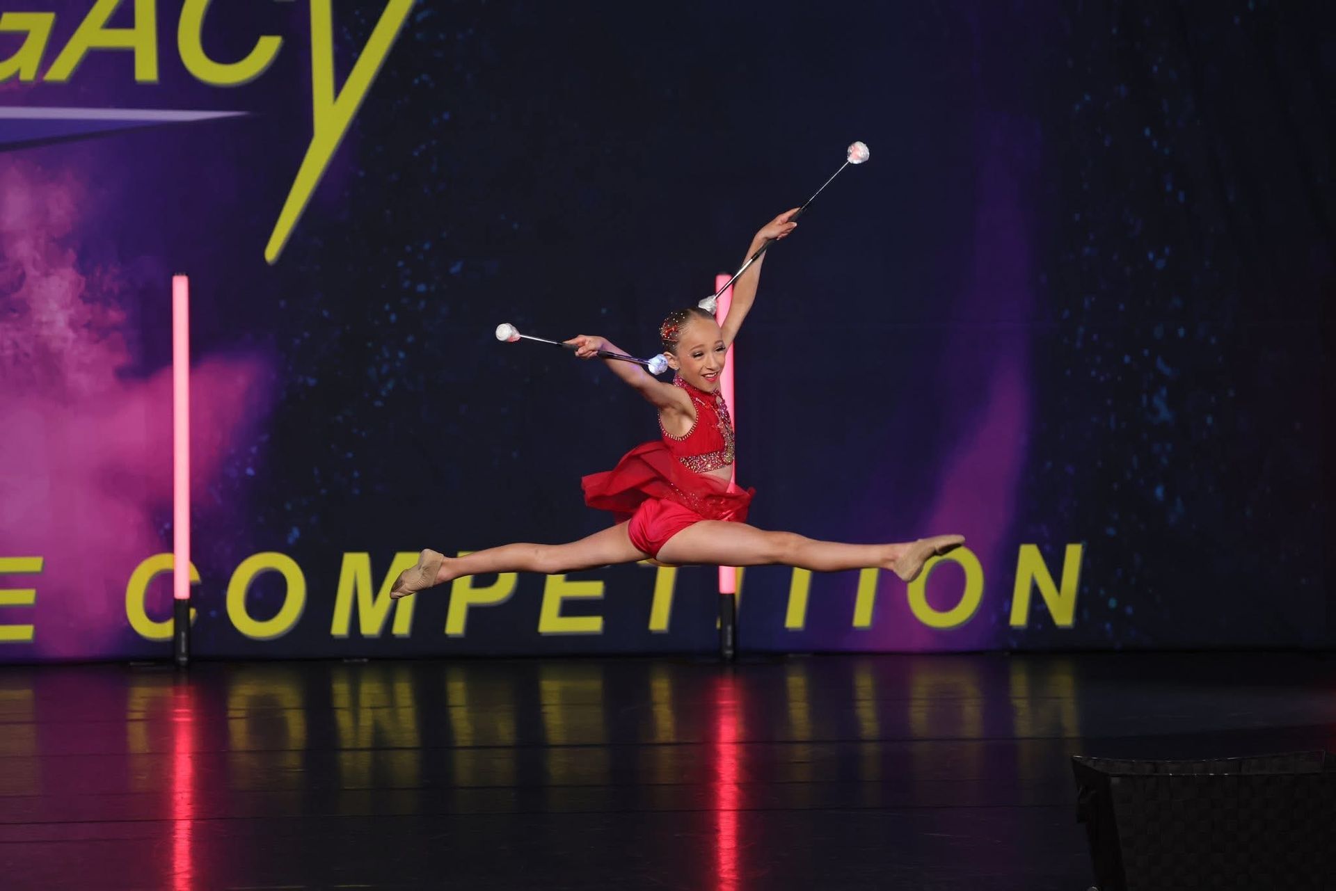 Young baton twirler in a red costume does a split in mid-air on stage.