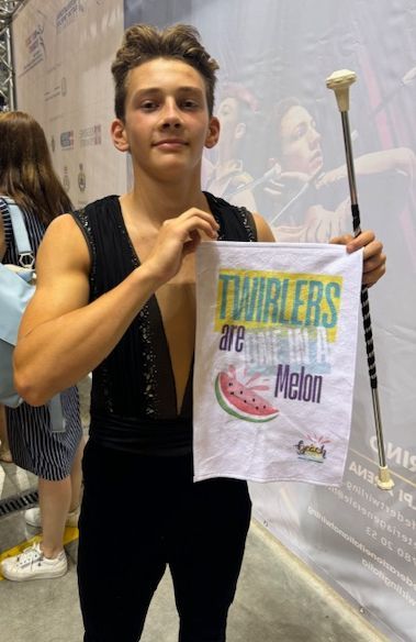 Young person in black outfit holding a sign, baton, and smiling. Sign reads: