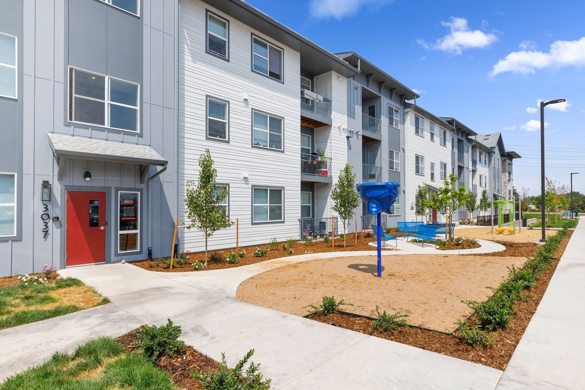 A modern multi-story apartment building with a red entrance door, white and grey exterior, and a sunny, landscaped yard.