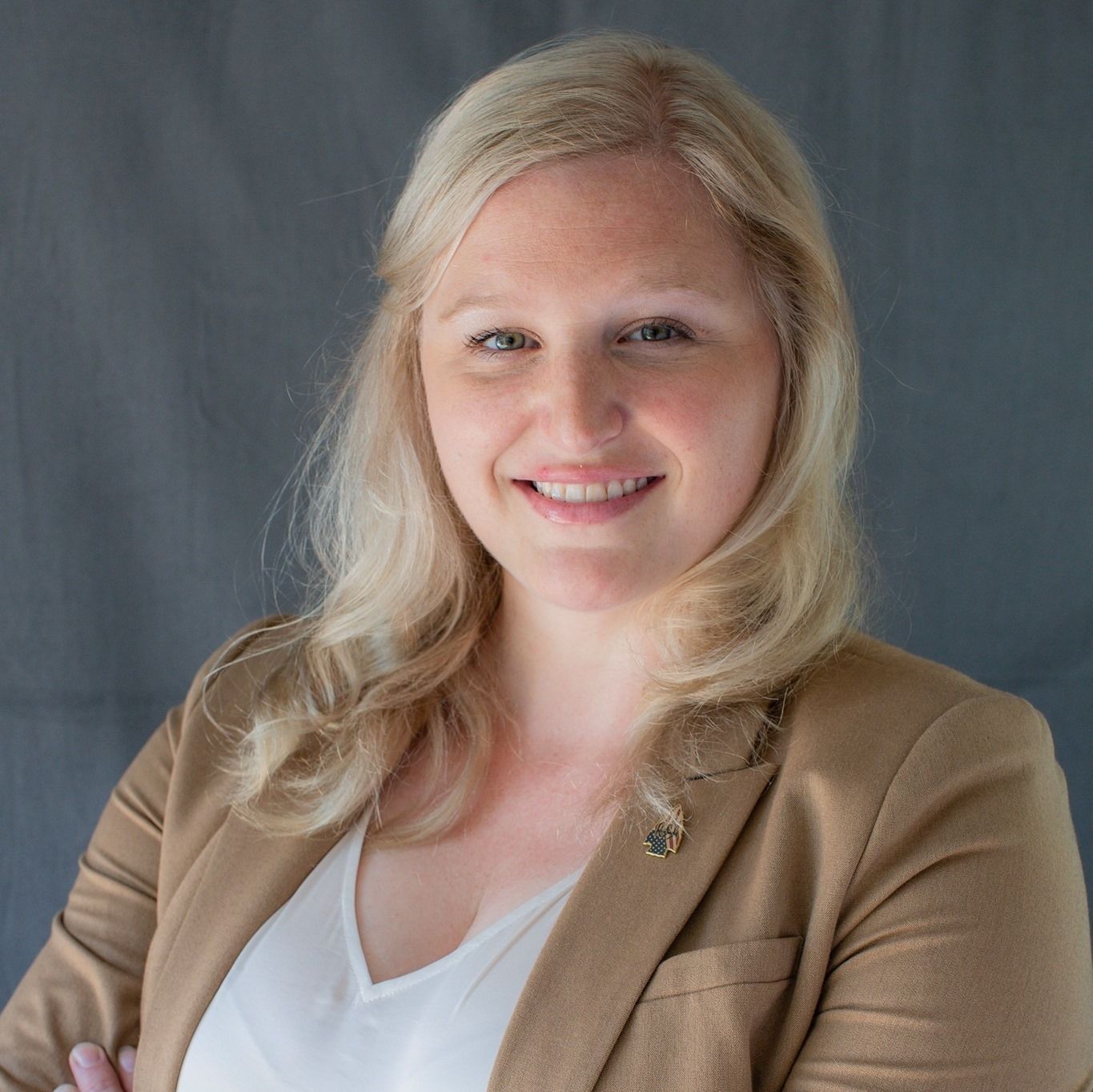 Blonde woman smiles, arms crossed, wearing a tan blazer and white shirt. Against a gray backdrop.
