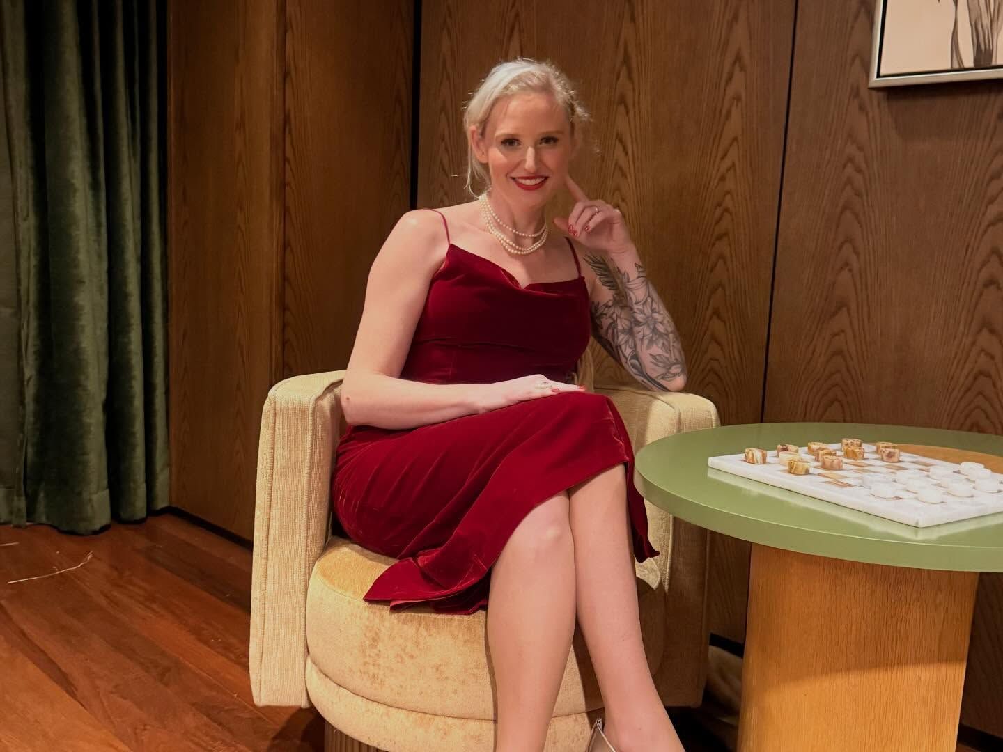 Woman in red dress sits in chair, arm tattoo visible, near small table, indoor setting.