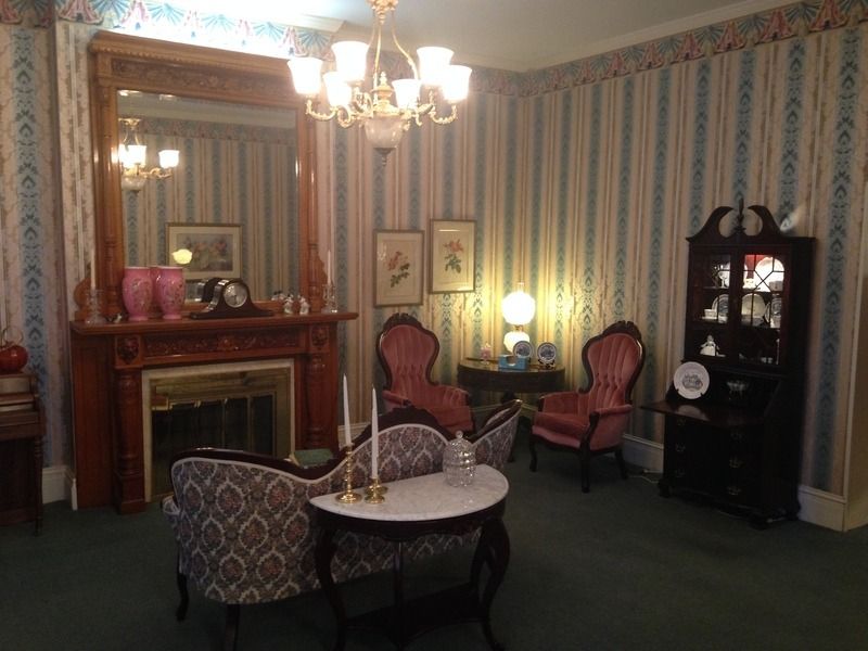 An antique parlor with patterned wallpaper, fireplace, vintage furniture, and a chandelier.