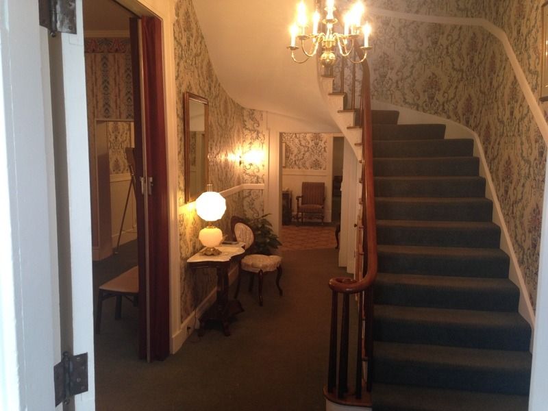 Interior view: Curved staircase with green carpet, wallpaper, ornate chandelier, and antique furniture.