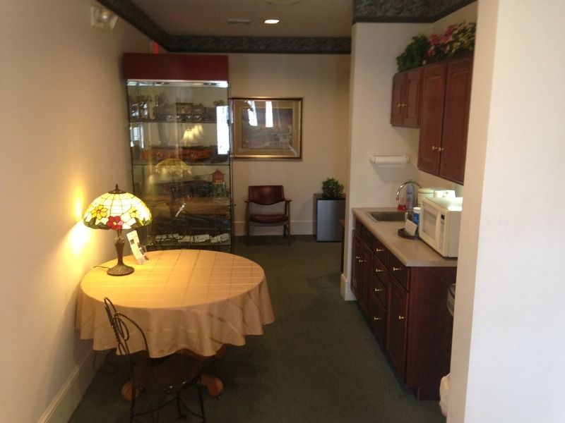 A small room with a round table, display cabinet, mini-kitchen, and seated chair.