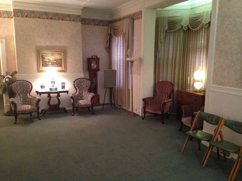 A funeral home viewing room with antique chairs, a grandfather clock, and a window with curtains.