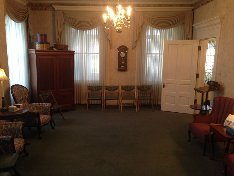 A dimly lit room with chairs, windows, and a chandelier. A tall cabinet is on the left.