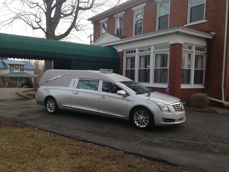 Silver hearse parked in front of a brick building with a covered entrance.